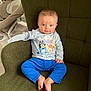 baby, child, blue_eyes, cartoon_shirt, blue_pants, green_chair, indoor, cute, sitting, barefoot, smiling, curious, young_child, soft_lighting, home_interior, comfortable, portrait, toddler, innocent, cozy