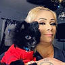 black_dog, blonde_hair, bracelet, ceiling_fan, curtains, disco_ball, dog, face, holding, indoor, living_room, portrait, puppy, red_sweater, selfie, smartwatch, smile, sparkle_effect, tattoo, woman