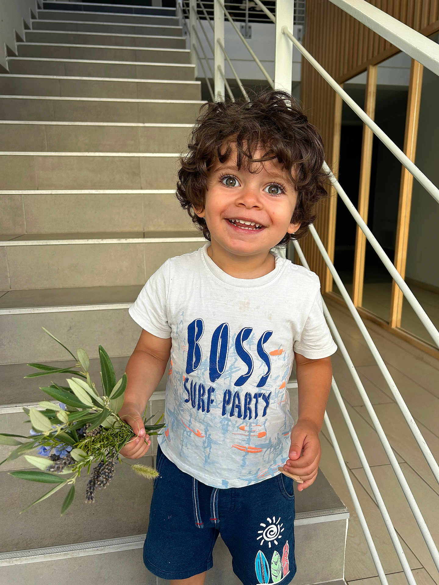 Nael participe au concours pour gagner de l'argent avec cette photo : bouquet, casual_clothing, child, curly_hair, cute, eyes, flowers, hair, happy, holding, indoor, person, portrait, railing, shorts, smiling, staircase, standing, toddler, tshirt
