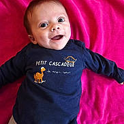 Lylio a rejoint le concours — aidez-le/la à gagner de superbes lots ! baby, infant, child, smiling, portrait, face, eyes, open_mouth, navy_shirt, pink_blanket, text_on_clothing, dinosaur_graphic, laying_down, happy, cheeks, short_hair, clothing, onesie, indoor, adorable