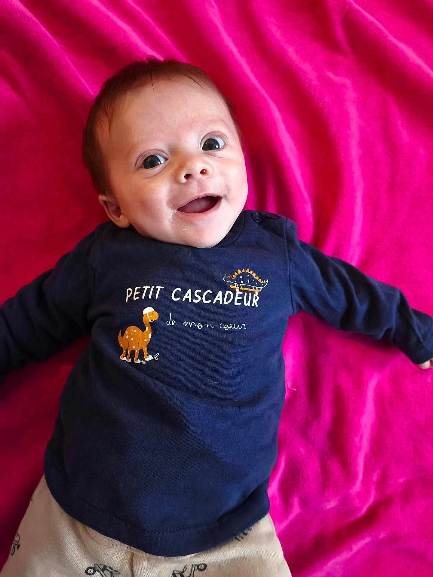 Lylio a rejoint le concours — aidez-le/la à gagner de superbes lots ! baby, infant, child, smiling, portrait, face, eyes, open_mouth, navy_shirt, pink_blanket, text_on_clothing, dinosaur_graphic, laying_down, happy, cheeks, short_hair, clothing, onesie, indoor, adorable