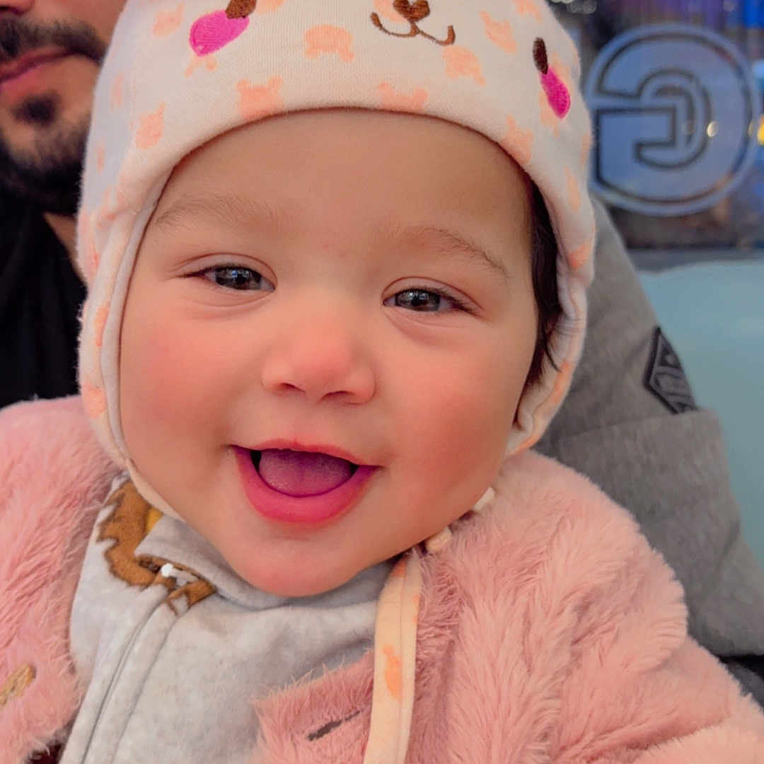 Saarah joined the competition — help win amazing prizes! apparel, baby, background, beard, bunny_ears, child, closeup, clothing, cute, face, happy, hat, indoor, infant, man, person, pink_jacket, portrait, smiling, warm