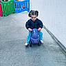 toddler, child, toy_car, purple, curly_hair, pigtails, jeans, jacket, sneakers, outdoor, playground, concrete, wall, barrier, colorful, happy, smiling, riding, person, daytime