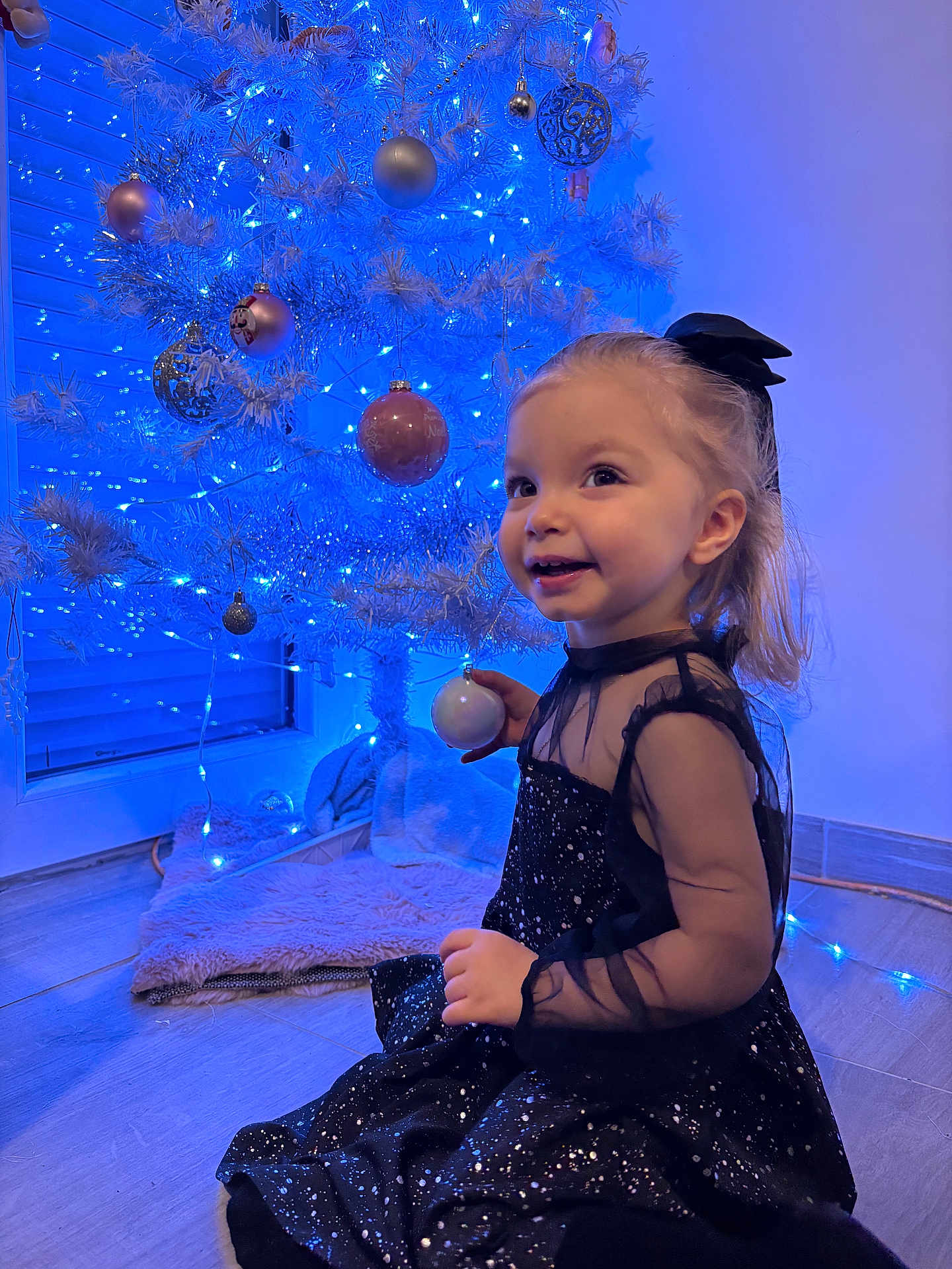 Luna participe au concours pour gagner de l'argent avec cette photo : child, toddler, christmas_tree, ornament, blue_lights, holiday, dress, smile, sitting, floor, decoration, bow, festive, indoor, portrait, sparkle, playful, tree_skirt, fairy_lights, wonder