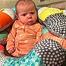 baby, infant, onesie, orange_clothing, soft_toys, cushions, patterned_fabric, baby_seat, indoor, child, cute, serious_expression, face, head, small_hands, plush, colorful, comfort, seated, young_child