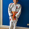 child, boy, judo_gi, orange_belt, martial_arts, barefoot, blue_wall, wooden_floor, standing, smiling, indoor, sport, uniform, young, portrait, casual, happy, person, clothing, pose