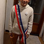 Soran participe au concours pour gagner de l'argent avec cette photo : child, boy, smile, sweater, pants, indoor, standing, sash, badge, wooden_door, floor, home, casual_clothing, happy, portrait, young, person, footwear, knitwear, button