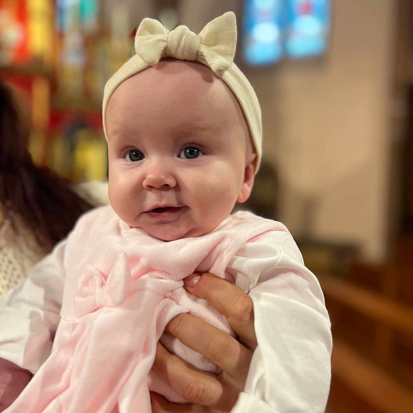 Scarlett participe au concours pour gagner de l'argent avec cette photo : baby, infant, face, headband, bow, pink_clothing, hand, person, portrait, indoors, blurred_background, stained_glass, church, cute, soft_light, child, holding, closeup, warm, smiling