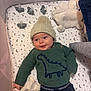 baby, infant, hat, knitted_hat, pom_pom, green_sweater, dinosaur_design, crib, pacifier, teddy_bear, plush_toy, diaper, blanket, moon_pattern, star_pattern, sleeping_area, child, cute, smiling, cozy