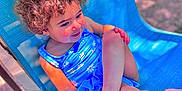 Ayla is registered to the contest to win money with this photo: child, toddler, curly_hair, blue_swimsuit, outdoor_chair, sunlight, relaxed, smiling, barefoot, summer, playful, skin, person, seat, leisure, daylight, happy, resting, blue, texture