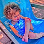 child, toddler, curly_hair, blue_swimsuit, outdoor_chair, sunlight, relaxed, smiling, barefoot, summer, playful, skin, person, seat, leisure, daylight, happy, resting, blue, texture