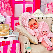 Ayla is registered to the contest to win money with this photo: baby, pink, robe, heart_shaped_sunglasses, armchair, pillow, shopping_bag, wooden_blocks, makeup_brush, pink_decor, toy, child, cute, sleeping, indoor, plush, fashion, accessory, relaxing, portrait