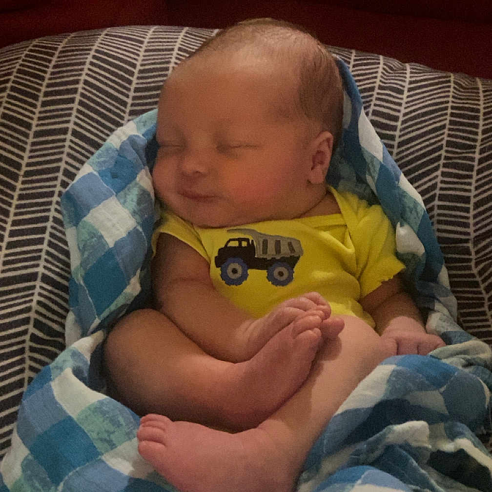 Creed joined the competition — help win amazing prizes! baby, blanket, blue, checkered, child, cute, dump_truck, face, feet, indoor, infant, onesie, patterned_cushion, peaceful, red_background, resting, sleeping, soft_lighting, toes, yellow_clothing