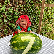 Beatrice joined the competition — help win amazing prizes! baby, child, watermelon, number_seven, fruit, table, checkered_tablecloth, greenery, ivy, outdoor, hat, red_clothing, smiling, happy, cute, nature, summer, garden, wooden_post, portrait