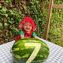 baby, child, watermelon, number_seven, fruit, table, checkered_tablecloth, greenery, ivy, outdoor, hat, red_clothing, smiling, happy, cute, nature, summer, garden, wooden_post, portrait