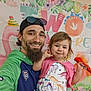 child, toddler, adult_male, beard, cap, sunglasses, smile, selfie, candy, dress, dinosaur_print, party_banner, floral, colorful, indoor, celebration, holding, portrait, happy, family