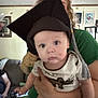 baby, graduation_cap, person, indoor, living_room, couch, hand, green_shirt, curly_hair, drool, face, portrait, child, clothing, furniture, photo_frame, wall, holding, blur, casual