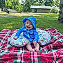 Weston is registered to the contest to win money with this photo: baby, child, blue_hat, blue_clothing, plaid_blanket, pillow, grass, park, trees, sunny, outdoor, smiling, happy, sitting, nature, daylight, infant, cute, playful, relaxing