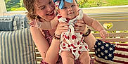 Genesis is registered to the contest to win money with this photo: baby, child, woman, smiling, porch, swing, pillow, american_flag, outdoor, daylight, greenery, tree, house, curly_hair, dress, bow, holding, happy, skin, candid