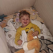 Chris a rejoint le concours — aidez-le/la à gagner de superbes lots ! bed, blanket, child, cozy, cute, face, furniture, happy, home, indoor, person, pillow, plush_toy, resting, sleepwear, smile, soft_toy, toddler, toy, yellow_sweater