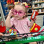toddler, shopping_cart, pink_sunglasses, toy, snack, store_interior, autumn_decor, flowers, child, baby, retail, colorful, hair_bows, clothing, hand, face, person, sitting, plastic_toys, supermarket