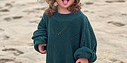 Esmée a rejoint le concours — aidez-le/la à gagner de superbes lots ! child, toddler, beach, sand, barefoot, sweater, green_sweater, curly_hair, tongue_out, smiling, playful, standing, outdoor, daylight, cute, young_child, fun, casual_clothing, nature, portrait
