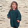 child, toddler, beach, sand, barefoot, sweater, green_sweater, curly_hair, tongue_out, smiling, playful, standing, outdoor, daylight, cute, young_child, fun, casual_clothing, nature, portrait