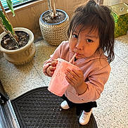 Valery joined the competition — help win amazing prizes! child, drink, pink, straw, indoor, potted_plant, mat, floor, window, blinds, shoes, jacket, curious, toddler, hair, hand, face, person, standing, beverage