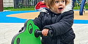 Mayllie participe au concours pour gagner de l'argent avec cette photo : child, toddler, playground, toy, rocker, green, dinosaur, jacket, black_jacket, blue_pants, outdoor, play, fun, happy, expression, fence, tree, grass, residential, daylight