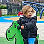 child, toddler, playground, toy, rocker, green, dinosaur, jacket, black_jacket, blue_pants, outdoor, play, fun, happy, expression, fence, tree, grass, residential, daylight