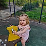 child, toddler, playground, yellow_rocker, outdoor, fence, greenery, grass, pathway, summer_clothing, pink_dress, white_sandals, hair_clip, playing, park, daylight, expression, young_kid, fun, recreation