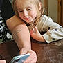 child, girl, adult, arm, smartphone, table, hair_scrunchie, tattoo, indoor, curious, leaning, wooden_table, long_sleeve, polka_dot, candid, family, technology, screen, hand, furniture