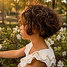 child, side_profile, curly_hair, golden_hour, sunlight, flower, white_flower, outdoor, nature, park, greenery, portrait, summer, daylight, young, person, dress, white_dress, beautiful, peaceful