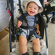 James is registered to the contest to win money with this photo: toddler, child, baby, smiling, hat, bucket_hat, stroller, straps, plaid_shorts, blue_shirt, boots, indoor, wooden_floor, baby_gear, happy, sitting, person, face, footwear, clothing