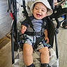 toddler, child, baby, smiling, hat, bucket_hat, stroller, straps, plaid_shorts, blue_shirt, boots, indoor, wooden_floor, baby_gear, happy, sitting, person, face, footwear, clothing