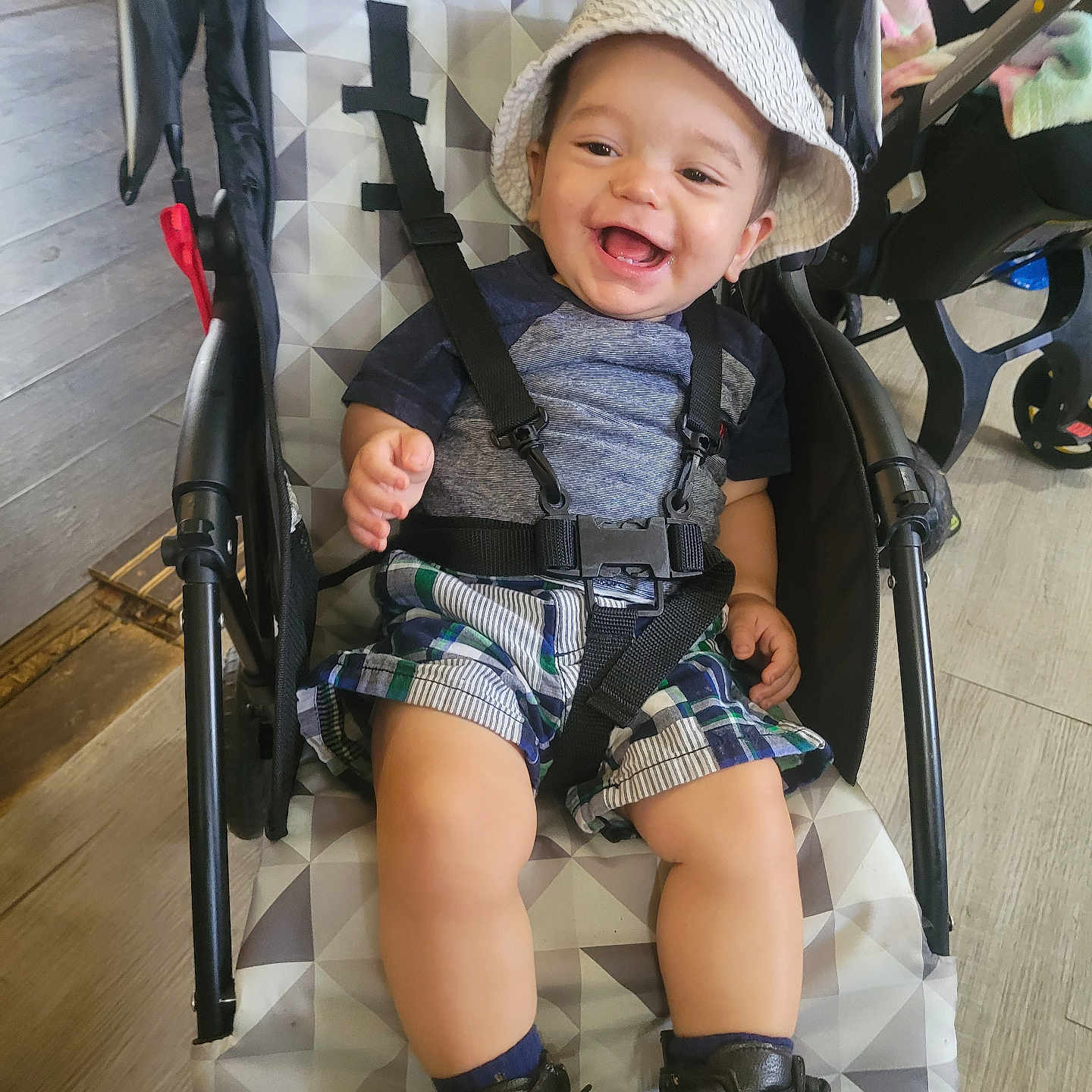 James is registered to the contest to win money with this photo: toddler, child, baby, smiling, hat, bucket_hat, stroller, straps, plaid_shorts, blue_shirt, boots, indoor, wooden_floor, baby_gear, happy, sitting, person, face, footwear, clothing
