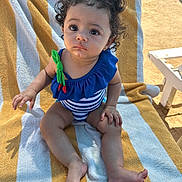 Mikayla joined the competition — help win amazing prizes! toddler, child, beach_towel, swimsuit, curly_hair, blue_swimsuit, striped_towel, outdoor, sunny, barefoot, sitting, cute, expression, young_child, summer, playful, vacation, sand, daylight, person