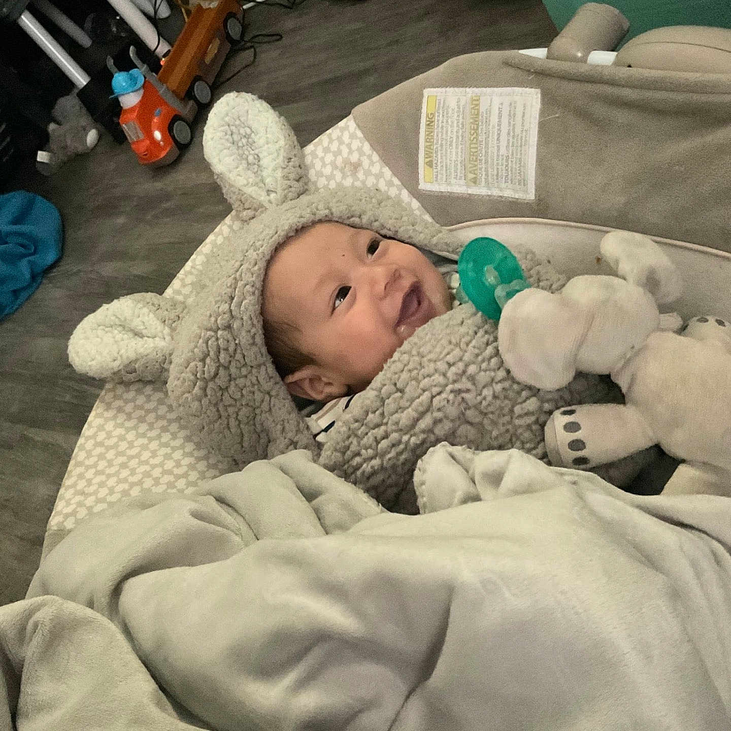 Micheal joined the competition — help win amazing prizes! baby, blanket, child, cozy, cute, elephant, face, floor, happy, headwear, hoodie, indoor, infant, pacifier, plush_toy, smiling, soft_texture, toy_truck, warm_clothing, wooden_floor