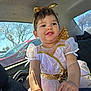 child, toddler, costume, dress, gold_bow, angel_wings, car_interior, steering_wheel, smile, portrait, window, sunlight, trees, earrings, hair, hands, seat, fabric, cute, happy