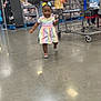 Kaylani is registered to the contest to win money with this photo: toddler, child, shopping_cart, store, aisle, dress, bows, shoes, floor, person, retail, shopping, happy, walking, colorful, clothing, shelves, product, indoor, grocery