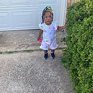 Kaylani joined the competition — help win amazing prizes! toddler, child, outdoor, driveway, bush, greenery, pineapple_pattern, shorts, tshirt, black_sneakers, beaded_hair, pink_toy, curious, standing, concrete, garage_door, brick_wall, casual_clothing, daylight, portrait