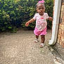 toddler, child, pink_clothing, bow, sandals, concrete, bushes, brick_wall, downspout, outdoor, sidewalk, plant, greenery, person, walking, cute, summer, sunny, young_child, expression
