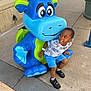 child, boy, statue, dinosaur, blue, green, sidewalk, smile, shorts, shirt, outdoor, concrete, playful, happy, casual_clothing, footwear, urban, daylight, person, toy