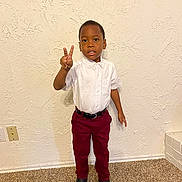 Reese is registered to the contest to win money with this photo: child, boy, peace_sign, white_shirt, burgundy_pants, black_shoes, standing, indoor, carpet, wall, textured_wall, expression, looking_at_camera, casual, portrait, single_person, young, curious, confident, floor