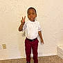 child, boy, peace_sign, white_shirt, burgundy_pants, black_shoes, standing, indoor, carpet, wall, textured_wall, expression, looking_at_camera, casual, portrait, single_person, young, curious, confident, floor