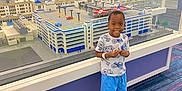 Reese is registered to the contest to win money with this photo: child, boy, lego, building_model, display_case, indoor, smile, carpet, clothing, shorts, shoes, glass, reflection, toy, architecture, exhibit, hand, standing, happy, person