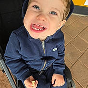 Gabriel is registered to the contest to win money with this photo: child, toddler, smiling, blue_hoodie, stroller, snack, teeth, face, happy, sitting, clothing, person, outdoor, pavement, tile_floor, hooded_jacket, hand, casual_wear, cute, portrait