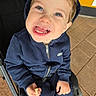 child, toddler, smiling, blue_hoodie, stroller, snack, teeth, face, happy, sitting, clothing, person, outdoor, pavement, tile_floor, hooded_jacket, hand, casual_wear, cute, portrait