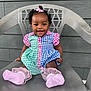 Séyori is registered to the contest to win money with this photo: baby, child, smiling, sitting, dress, pink_bow, pink_socks, chair, plastic_chair, outdoor, happy, cute, portrait, person, colorful_clothing, checkered_pattern, curly_hair, single_child, wall, feet