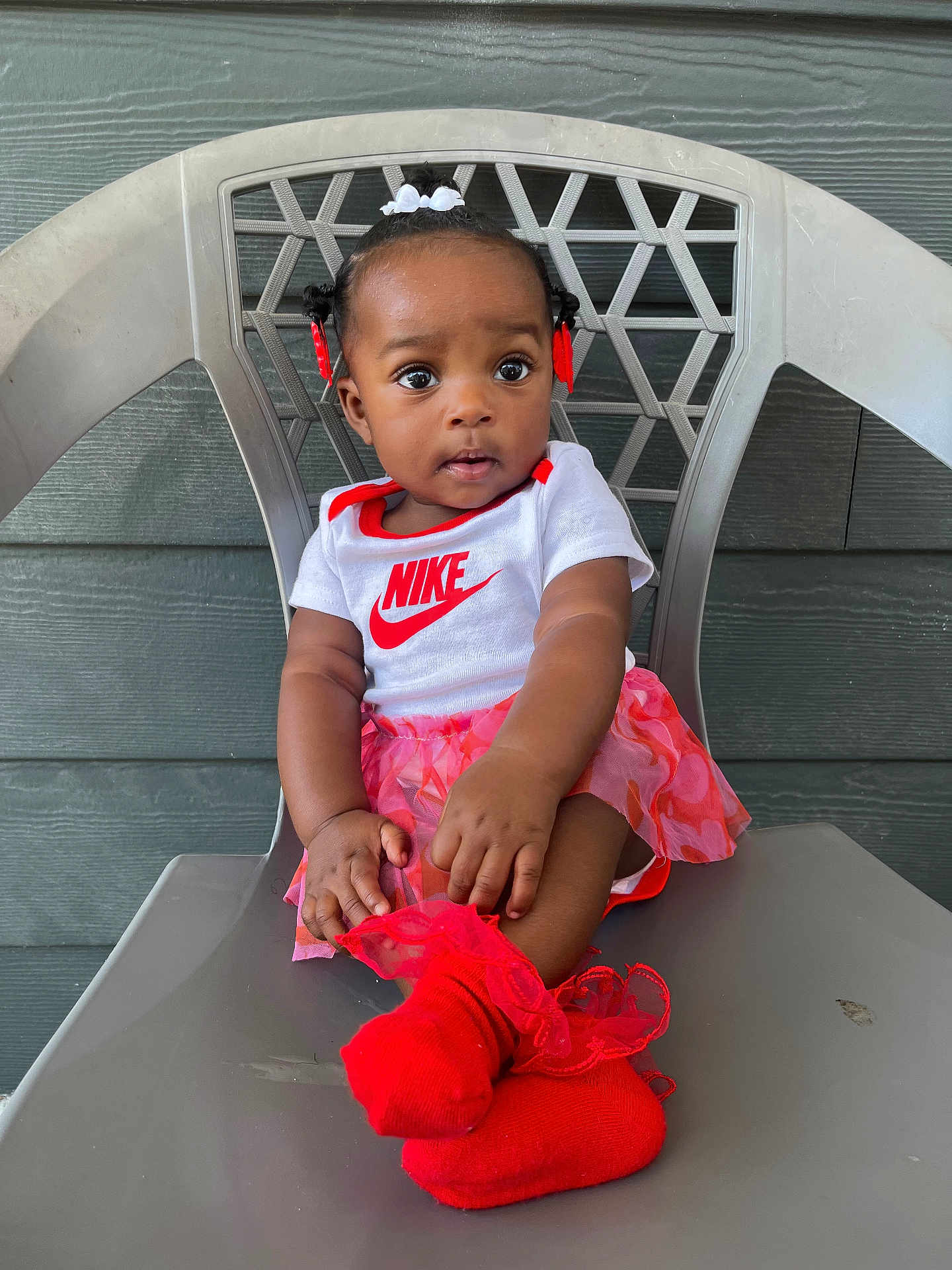 Séyori is registered to the contest to win money with this photo: baby, child, girl, sitting, chair, plastic_chair, clothing, onesie, socks, red_socks, skirt, hair_bows, face, eyes, hands, outdoor, wall, expression, nike_logo, cute